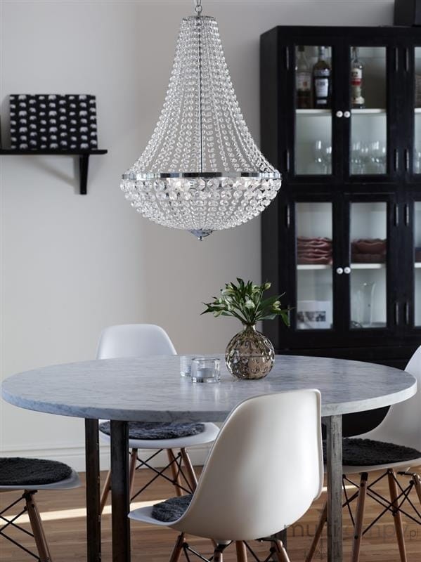 Lampa wisząca Granso 3-punktowa w kolorze chromu, wykonana z przezroczystych kryształów, zawieszona nad okrągłym stołem ze szklanym blatem. W tle czarna witryna z naczyniami i rośliną na stole.