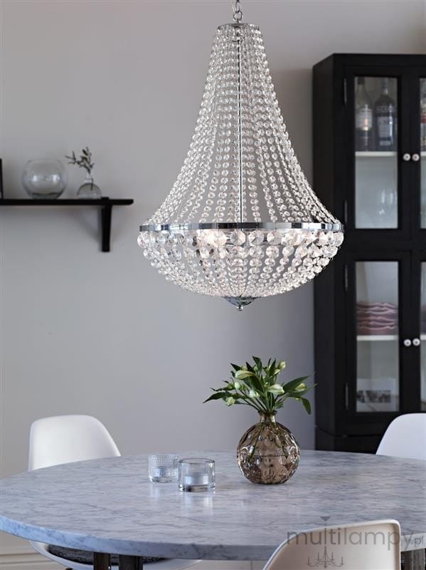 Lampa wisząca Granso 3-punktowa w chromie, o kształcie stożkowym, pokryta przezroczystymi kryształkami. Zawieszona nad okrągłym stołem z marmurowym blatem, w otoczeniu szklanych naczyń i wazonu z zielonymi roślinami.