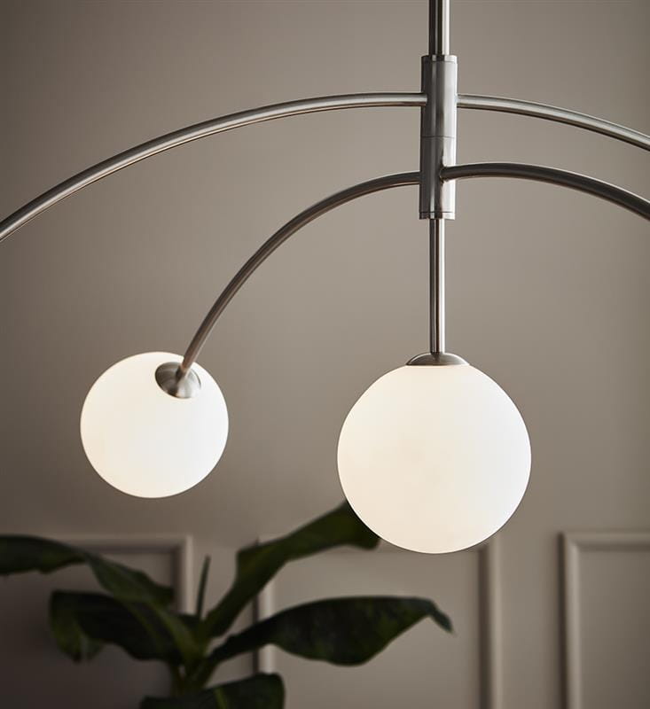 Lampa wisząca Heaven 5-punktowa w stalowej oprawie z dwoma białymi, okrągłymi kloszami. Ujęcie z góry, podkreślające delikatne łuki ramion. Tło w stonowanej kolorystyce, z widoczna rośliną w dolnym rogu.
