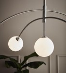 Lampa wisząca Heaven 5-punktowa w stalowej oprawie z dwoma białymi, okrągłymi kloszami. Ujęcie z góry, podkreślające delikatne łuki ramion. Tło w stonowanej kolorystyce, z widoczna rośliną w dolnym rogu.