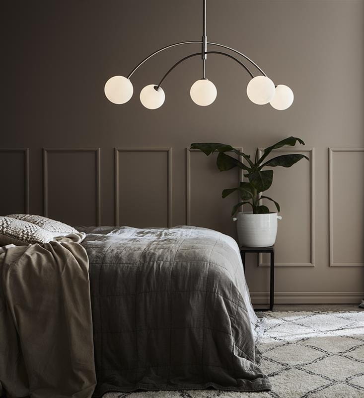 Lampa wisząca Heaven 5-punktowa w stalowej konstrukcji, z pięcioma okrągłymi, mlecznymi kloszami. Zawieszona nad łóżkiem w sypialni, w otoczeniu rośliny w białym doniczce, na tle jasnej ściany z panelami.