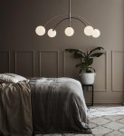 Lampa wisząca Heaven 5-punktowa w stalowej konstrukcji, z pięcioma okrągłymi, mlecznymi kloszami. Zawieszona nad łóżkiem w sypialni, w otoczeniu rośliny w białym doniczce, na tle jasnej ściany z panelami.