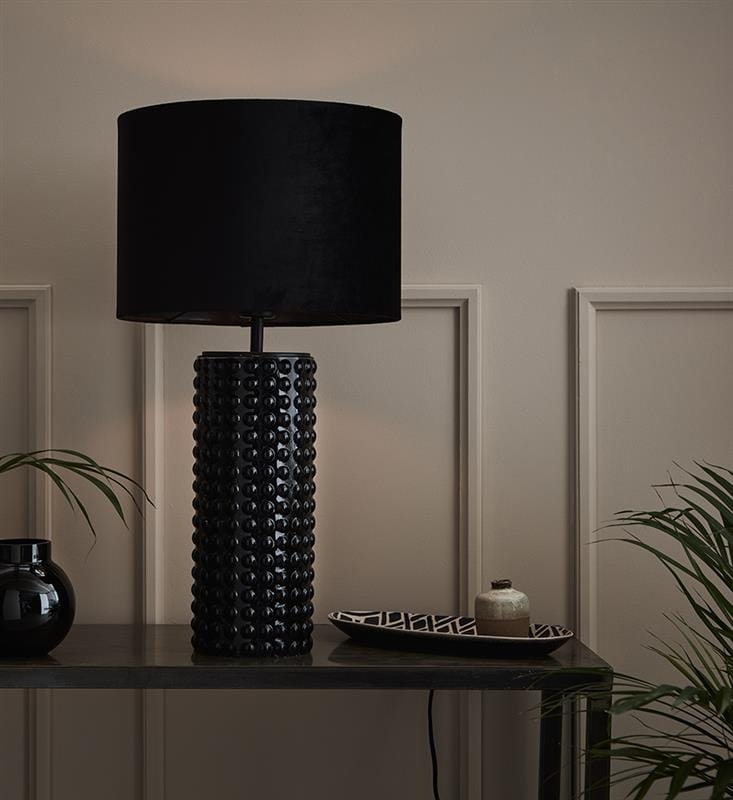 Lampa stołowa Proud w kolorze czarnym, z cylindrycznym, teksturowanym korpusem i aksamitnym kloszem. Ustawiona na ciemnym stole, obok dekoracyjnego talerza i doniczki, w tle gładka ściana.
