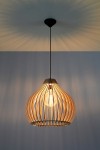 Lampa wisząca Aprilla SL.0639, 1-punktowa, o organicznym kształcie z drewna, w kolorze naturalnym. Zawieszona na cienkim czarnym kablu, z widoczną żarówką wewnątrz. Ujęcie frontalne, tło w odcieniu niebieskim, podkreślającym fakturę drewna.
