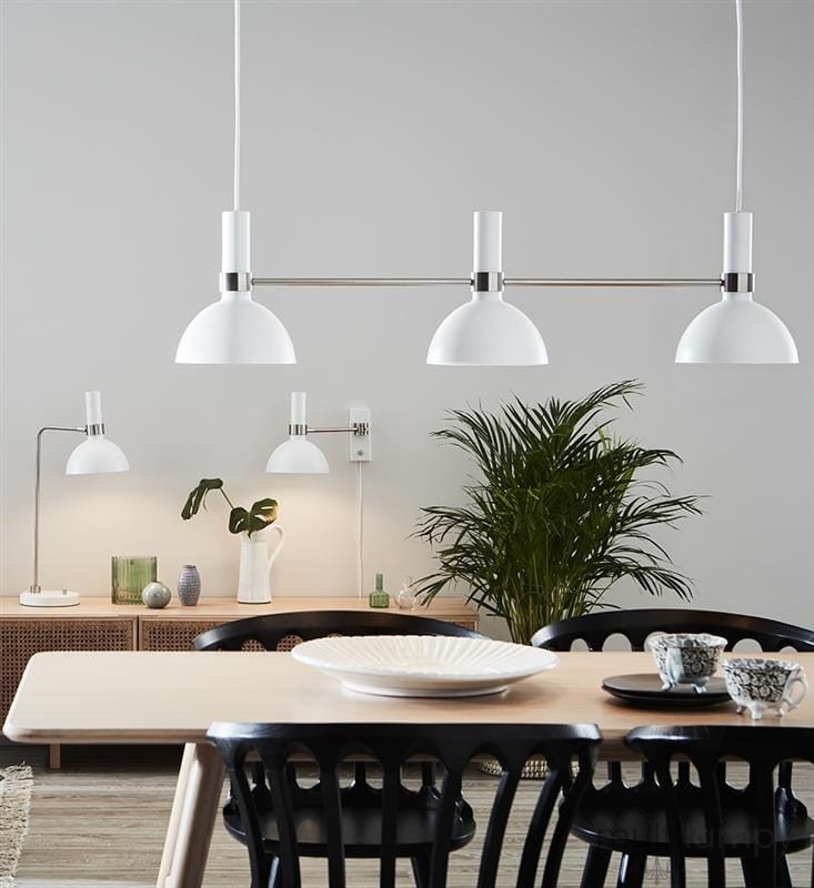 Lampa wisząca Larry 3-punktowa w białym kolorze z metalowymi elementami, zawieszona nad stołem. W tle widoczna roślina, a obok mniejsze lampy i dekoracje. Ujęcie frontalne, na jednolitym tle.