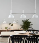 Lampa wisząca Larry 3-punktowa w białym kolorze z metalowymi elementami, zawieszona nad stołem. W tle widoczna roślina, a obok mniejsze lampy i dekoracje. Ujęcie frontalne, na jednolitym tle.