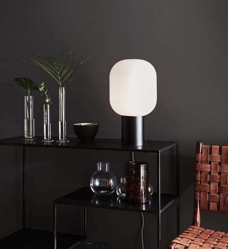 Lampa stołowa Brooklyn w białym kolorze z owalnym kloszem, umieszczona na czarnej półce. Obok znajdują się szklane wazoniki z roślinami oraz czarna miska. Tło w ciemnym odcieniu podkreśla nowoczesny design lampy.