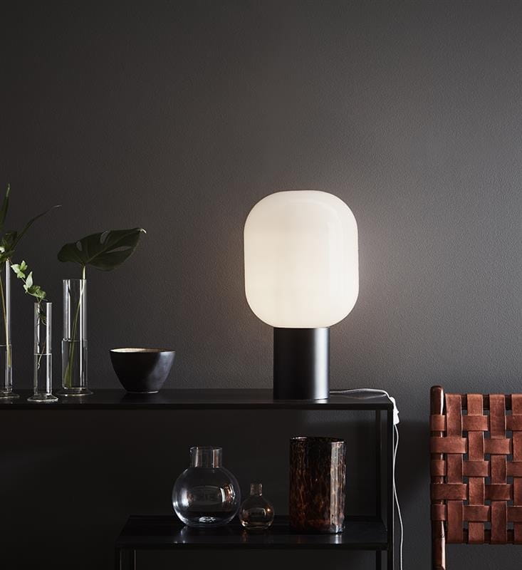 Lampa stołowa Brooklyn 1-punktowa w białej obudowie z okrągłym kloszem i czarną podstawą, ustawiona na ciemnym stoliku. W tle widoczne dekoracyjne szklane naczynia oraz rośliny w wazonach.