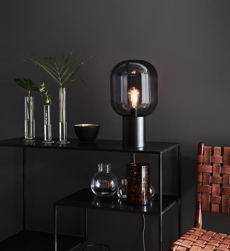 Lampa stołowa Brooklyn 1-punktowa z przydymionym szkłem, w kształcie okrągłym, na czarnej podstawie. Ustawiona na czarnej półce obok przezroczystych wazonów i ceramicznej miski. Tło w ciemnym kolorze podkreśla elegancki design.