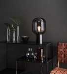 Lampa stołowa Brooklyn 1-punktowa z przydymionym szkłem, w kształcie okrągłym, na czarnej podstawie. Ustawiona na czarnej półce obok przezroczystych wazonów i ceramicznej miski. Tło w ciemnym kolorze podkreśla elegancki design.