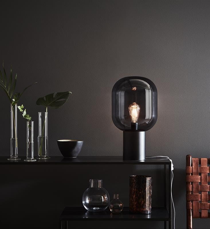 Lampa stołowa Brooklyn 1-punktowa, przydymiona, z okrągłym kloszem i czarną podstawą. Widoczna żarówka, elegancko eksponowana. Na tle ciemnej ściany, obok dekoracyjne elementy, w tym wazoniki i miseczka.