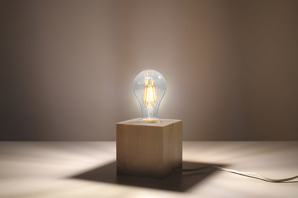 Lampka stołowa Ariz Drewno z przezroczystą żarówką na drewnianym, kwadratowym podstawie. Ujęcie frontalne, produkt oświetlający stół, widoczny kabel. Tło w odcieniach beżu, podkreślające nowoczesny design.