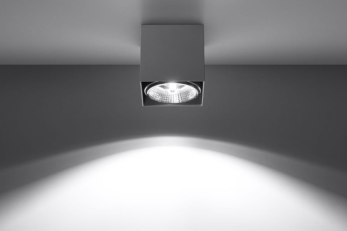 Lampa sufitowa Blake 1-punktowa w kolorze szarym, o kwadratowej podstawie. Widoczna lampa z reflektorem w centralnej części, emitująca światło na boki. Zamontowana na suficie, na tle gładkiej, jasnej powierzchni.