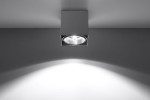 Lampa sufitowa Blake 1-punktowa w kolorze szarym, o kwadratowej podstawie. Widoczna lampa z reflektorem w centralnej części, emitująca światło na boki. Zamontowana na suficie, na tle gładkiej, jasnej powierzchni.