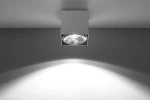 Lampa sufitowa Blake 1-punktowa w kolorze białym, o kwadratowym kształcie. Widoczna osłona z przezroczystym kloszem, emitującym światło w dół. Zainstalowana na suficie, na tle gładkiej, szarej powierzchni.