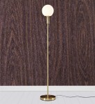 Lampa podłogowa Minna w kolorze mosiądzu z okrągłą, białą kloszem. Stojąca, z cienką nogą i okrągłą podstawą. Ujęcie frontalne na tle ciemnego drewna. Kabel zasilający widoczny u podstawy.