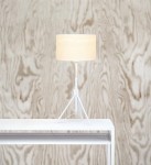 Biała lampka stołowa Sling z jedną żarówką, z cylindrycznym kloszem i trójnogą podstawą. Ustawiona na białym stole, w tle widoczna struktura drewniana. Prosta forma, idealna do nowoczesnych wnętrz.