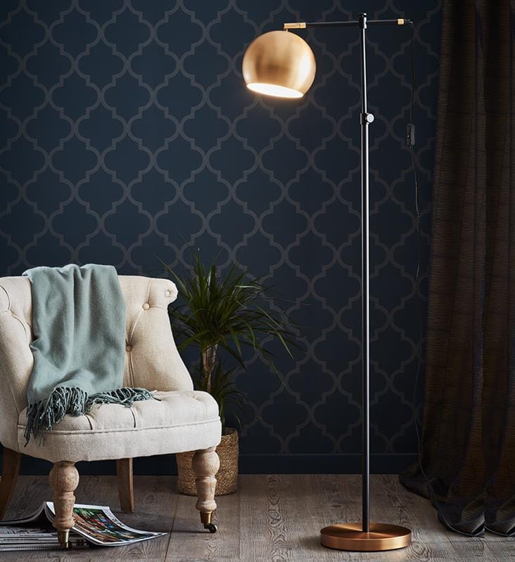 Lampa podłogowa Chester 1-punktowa w kolorze czarnym i brązowym, z okrągłym, złotym kloszem. Ustawiona obok jasnej, wygodnej sofy z niebieskim kocem. Tło w ciemnym kolorze z dekoracyjnym wzorem. Stojąca na okrągłej podstawie.