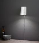 Biały kinkiet Barcelona 1-punktowy z okrągłym kloszem, zamontowany na szarej ścianie. Kabel zasilający biegnie w dół. Obok znajduje się czarna miska w białe wzory i butelka oliwy.