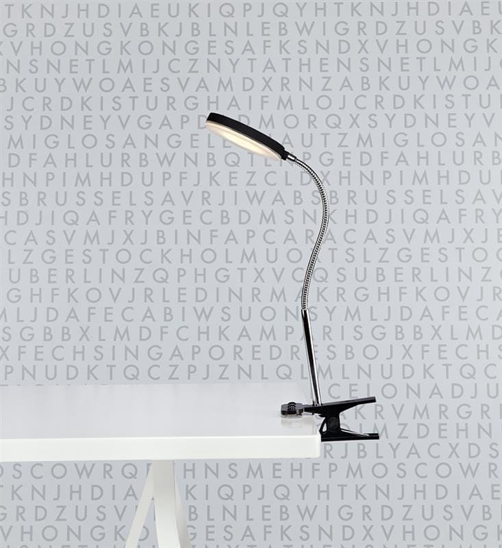 Flex Klip LED kinkiet 1-punktowy w kolorze czarnym i chromowanym, z elastycznym ramieniem, zamontowany na białym stole. Źródło światła umieszczone w wąskiej, prostokątnej obudowie. Tło z literowymi wzorami w odcieniach szarości.