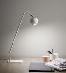 Lampka stołowa Coco w kolorze białym, z długim, zakrzywionym ramieniem i okrągłą głowicą. Ustawiona na prostokątnej podstawie, obok zeszytu, drewnianego pojemnika i kubka z długopisami. Tło jasnoszare, minimalistyczne.