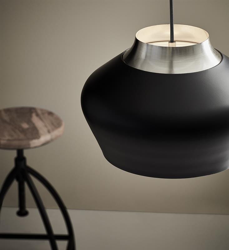 Lampa wisząca Kom D31 w kolorze czarnym z metalowym, stalowym wykończeniem. Posiada zaokrąglony kształt z matowej powierzchni. Wisi na cienkim przewodzie, z widocznym wnętrzem w neutralnym kolorze. W tle drewniane siedzisko na metalowej podstawie.