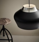 Lampa wisząca Kom D31 w kolorze czarnym z metalowym, stalowym wykończeniem. Posiada zaokrąglony kształt z matowej powierzchni. Wisi na cienkim przewodzie, z widocznym wnętrzem w neutralnym kolorze. W tle drewniane siedzisko na metalowej podstawie.