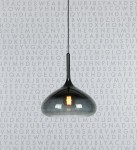Czarna lampa wisząca Cooper o jednopunktowym mocowaniu, o zaokrąglonym kształcie i połyskującej, szklanej obudowie. Zawieszona na cienkim kablu, z widoczną żarówką w środku, na jasnym tle z literami w tle.