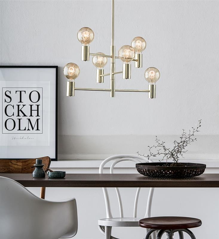 Lampa wisząca Capital 6-punktowa w złotym kolorze, z sześcioma okrągłymi kloszami na metalowych ramionach. Zawieszona nad drewnianym stołem, w tle plakat z napisem STOCKHOLM. Elementy dekoracyjne na stole, jasne tło.
