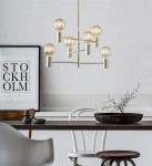 Lampa wisząca Capital 6-punktowa w złotym kolorze, z sześcioma okrągłymi kloszami na metalowych ramionach. Zawieszona nad drewnianym stołem, w tle plakat z napisem STOCKHOLM. Elementy dekoracyjne na stole, jasne tło.