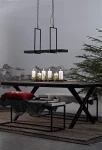 Czarna lampa wisząca Tray LED 3-punktowa, zawieszona na łańcuchach, umieszczona nad drewnianym stołem. W tle widoczna ściana z odrapanym tynkiem oraz dekoracje, takie jak świeczniki z białymi świecami i zielona choinka.