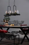 Lampa wisząca Tray LED w kolorze czarnym, 3-punktowa, z białymi świecami otoczonymi zielonymi gałązkami. Zawieszona na łańcuchu, umieszczona nad drewnianym stołem z owocami i dekoracjami, w otoczeniu czerwonych poduszek.