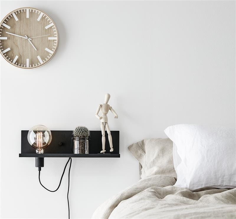 Kinkiet 1-punktowy w kolorze czarnym z portem USB, zamontowany na ścianie. Na półce znajdują się przezroczysta żarówka, szklany słoik oraz drewniana figurka. Tło jasne, z zegarem ściennym nad półką. W pobliżu widoczna pościel w odcieniach beżu i bieli.