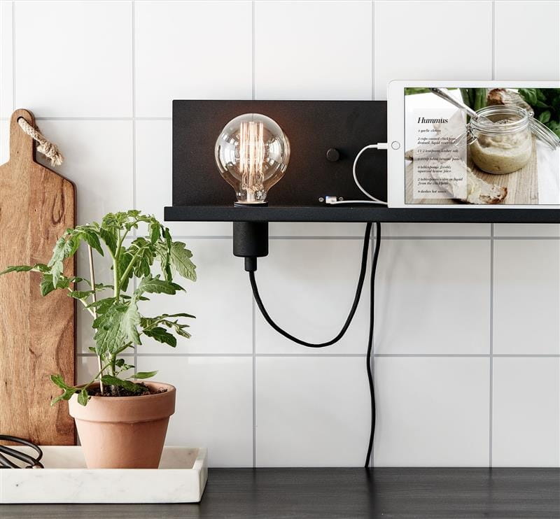 Kinkiet 1-punktowy Multi w kolorze czarnym z portem USB, umieszczony na półce. Oświetlenie z przezroczystą żarówką oraz widoczny kabel. W tle mała doniczka z rośliną i tablet z przepisem kulinarnym.