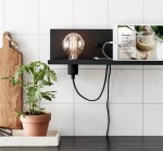 Kinkiet 1-punktowy Multi w kolorze czarnym z portem USB, umieszczony na półce. Oświetlenie z przezroczystą żarówką oraz widoczny kabel. W tle mała doniczka z rośliną i tablet z przepisem kulinarnym.