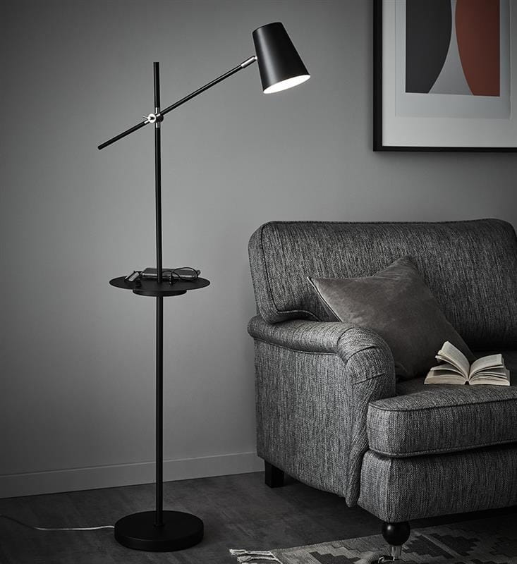Lampa podłogowa Hudson 1-punktowa w kolorze czarnym z regulowanym kloszem, zamontowana na cienkim stelażu. Posiada półkę na akcesoria. W tle szara sofa z poduszką i książką, na jednolitym, ciemnym tle.