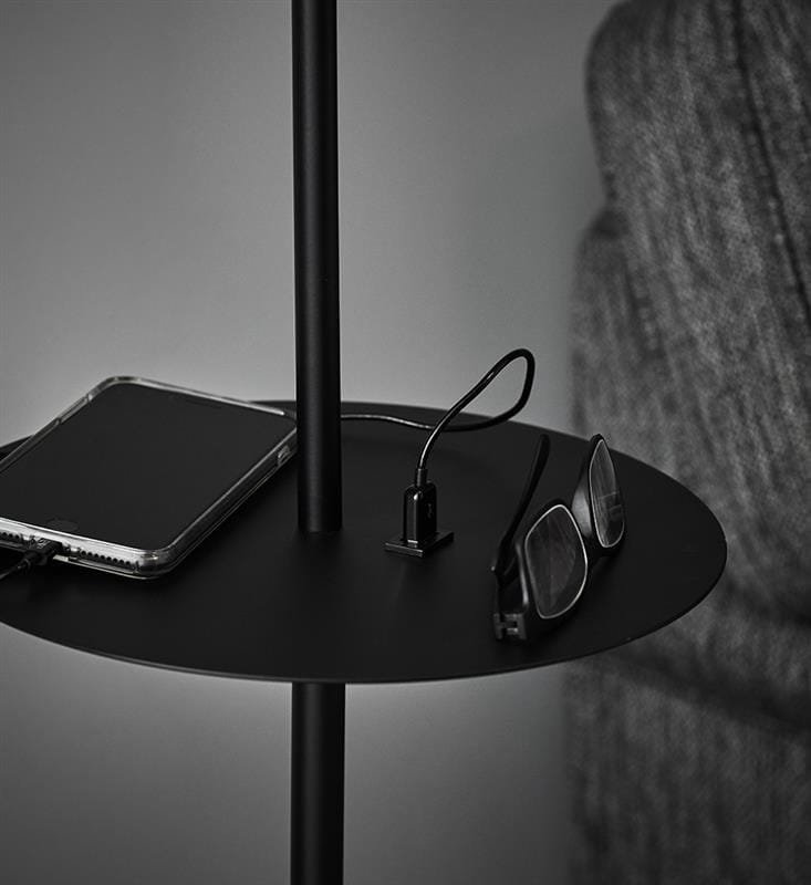 Czarna lampa podłogowa Hudson 1-punktowa z USB, z okrągłym, płaskim blatem. Na stole leży smartfon z przezroczystą obudową oraz okulary. Widoczny port USB do ładowania. Tło w odcieniach szarości, co podkreśla nowoczesny design lampy.