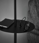 Czarna lampa podłogowa Hudson 1-punktowa z USB, z okrągłym, płaskim blatem. Na stole leży smartfon z przezroczystą obudową oraz okulary. Widoczny port USB do ładowania. Tło w odcieniach szarości, co podkreśla nowoczesny design lampy.