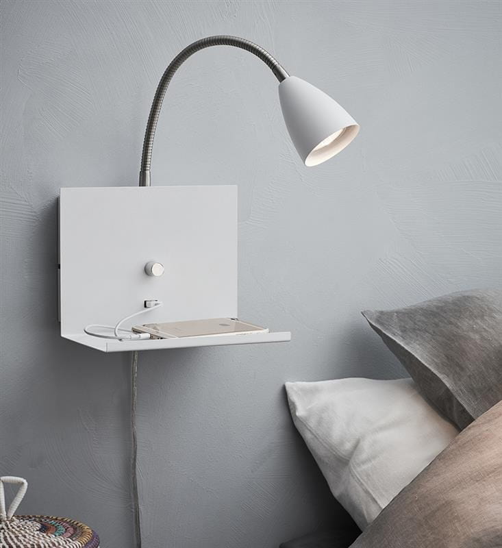 Kinkiet Logi w kolorze białym z elastycznym ramieniem, wyposażony w port USB. Produkt montowany na ścianie z małą półką na książki lub urządzenia elektroniczne, umieszczony obok poduszki na łóżku.