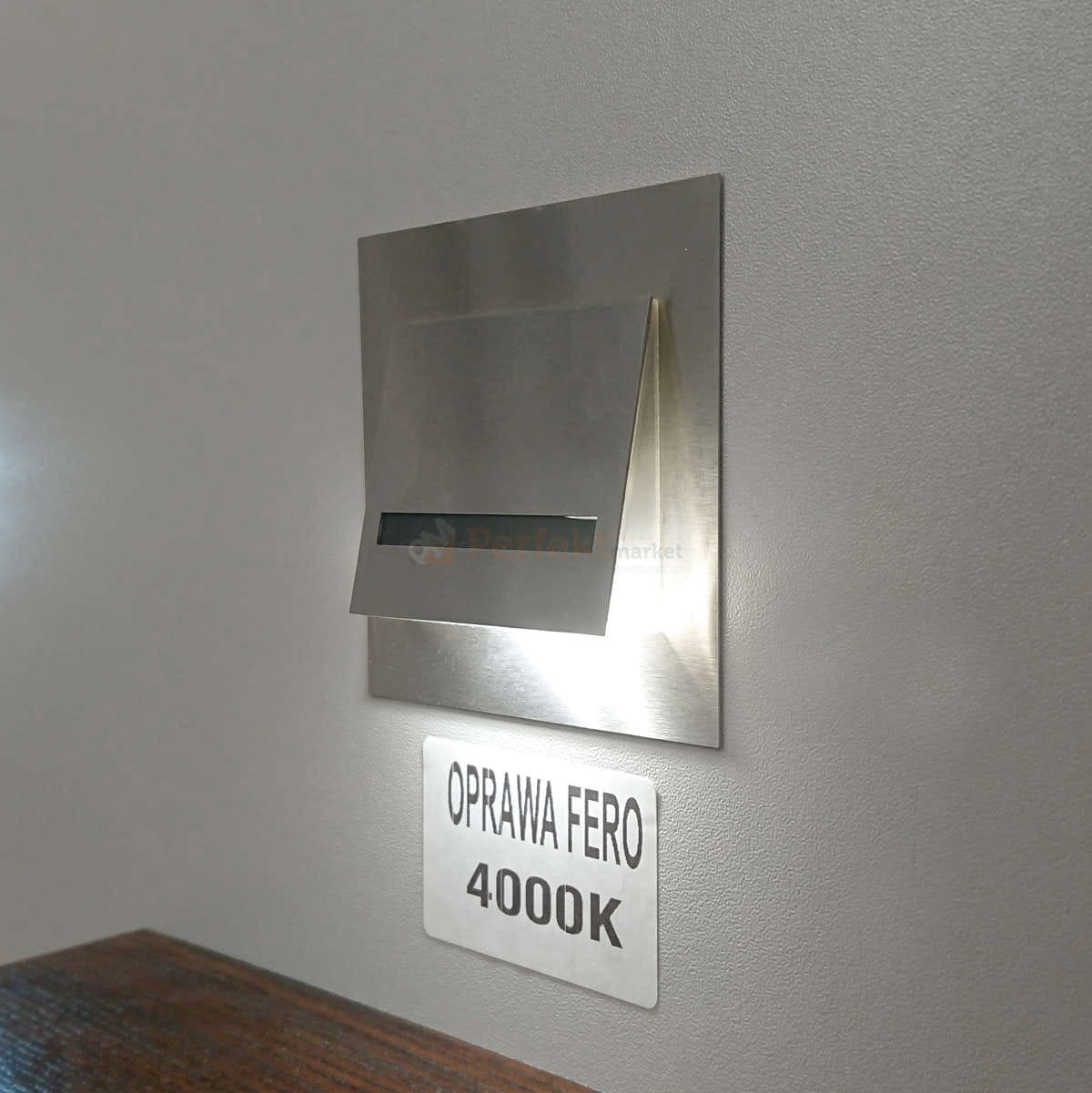 Oprawa schodowa Fero LED 230V w neutralnej barwie 4000K, wykonana ze stali nierdzewnej, montowana na ścianie. Posiada prostokątny kształt z wcięciem, emitującym światło w dół. Widoczne oznaczenie OPRAWA FERO 4000K na metalowej tabliczce.