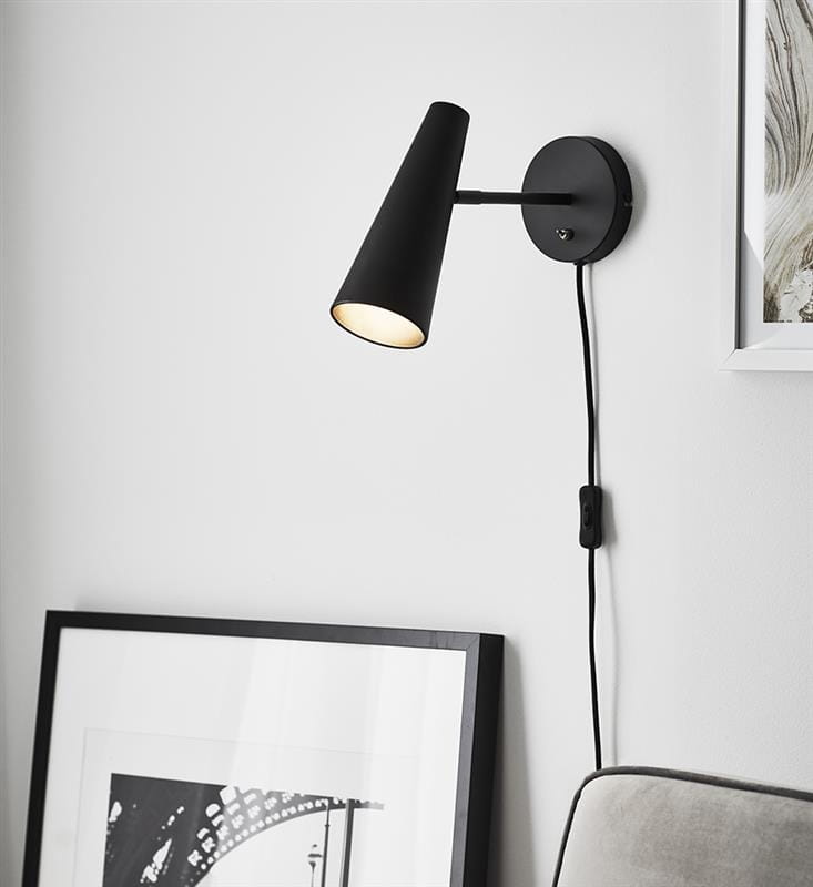 Kinkiet Peak 1-punktowy w kolorze czarnym, z cylindryczną podstawą i stożkowym kloszem. Ujęcie z boku, zamontowany na ścianie. Widać przewód z włącznikiem. W tle ramka z grafiką i część sofy w odcieniu szarości.