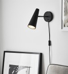 Kinkiet Peak 1-punktowy w kolorze czarnym, z cylindryczną podstawą i stożkowym kloszem. Ujęcie z boku, zamontowany na ścianie. Widać przewód z włącznikiem. W tle ramka z grafiką i część sofy w odcieniu szarości.