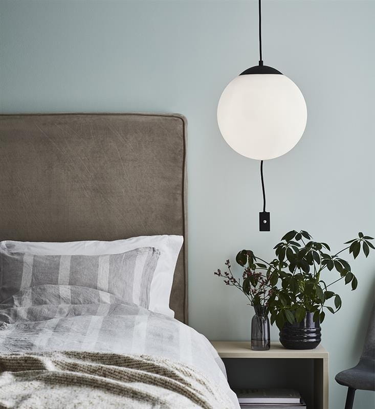 Lampa wisząca 3-punktowa Here w kształcie białej kuli, zawieszona nad łóżkiem. Czarny uchwyt i przewód. W tle szare wezgłowie, po lewej pościel w paski, a obok roślina w doniczce na stoliku nocnym. Ściana w jasnym kolorze.