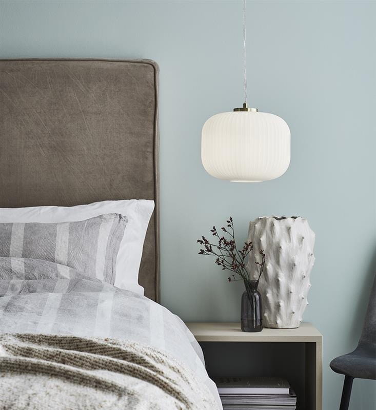 Lampa wisząca Sober 1-punktowa w kolorze białym z mosiężnym wykończeniem, o zaokrąglonym kształcie, zawieszona nad nocnym stolikiem. W tle szare łóżko z tekstylnym zagłówkiem oraz dekoracyjne wazon z gałązkami.