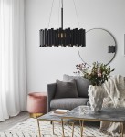 Lampa wisząca 3-punktowa w kolorze czarnym, z cylindrycznymi elementami, zawieszona na cienkim sznurku. W tle okrągłe lustro, szary fotel, stół z metalowymi nogami i dekoracje roślinne w wazonie. Wnętrze jasne z zasłonami.