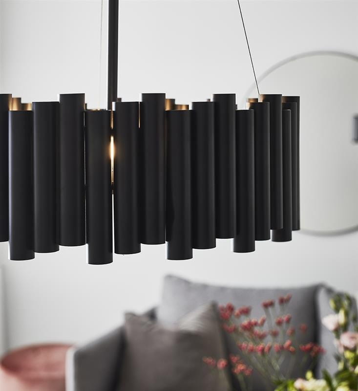 Czarna lampa wisząca 3-punktowa Level z cylindrycznymi elementami, zawieszona na cienkim przewodzie. W tle widoczna część wnętrza z szarymi poduszkami i dekoracyjnymi roślinami. Lampa ma elegancki, nowoczesny design.