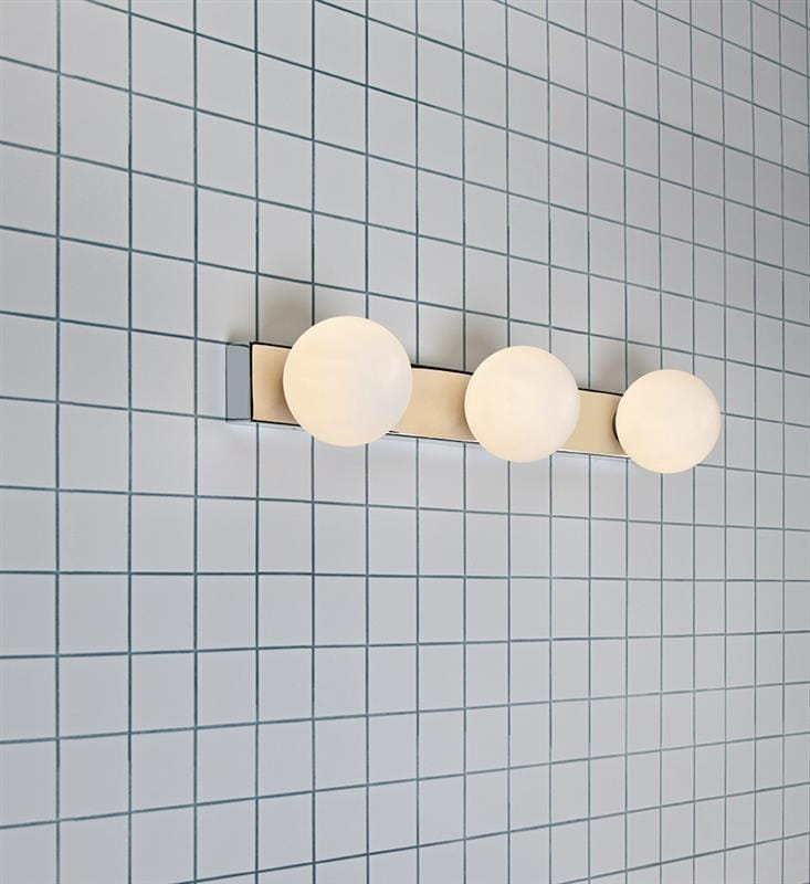 Mini kinkiet 3-punktowy w chromie z białymi, okrągłymi kloszami. Ułożony poziomo, montowany na ścianie w jasnym otoczeniu z niebieską, kafelkową powierzchnią. Klosze zapewniają równomierne oświetlenie.