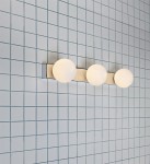 Mini kinkiet 3-punktowy w chromie z białymi, okrągłymi kloszami. Ułożony poziomo, montowany na ścianie w jasnym otoczeniu z niebieską, kafelkową powierzchnią. Klosze zapewniają równomierne oświetlenie.