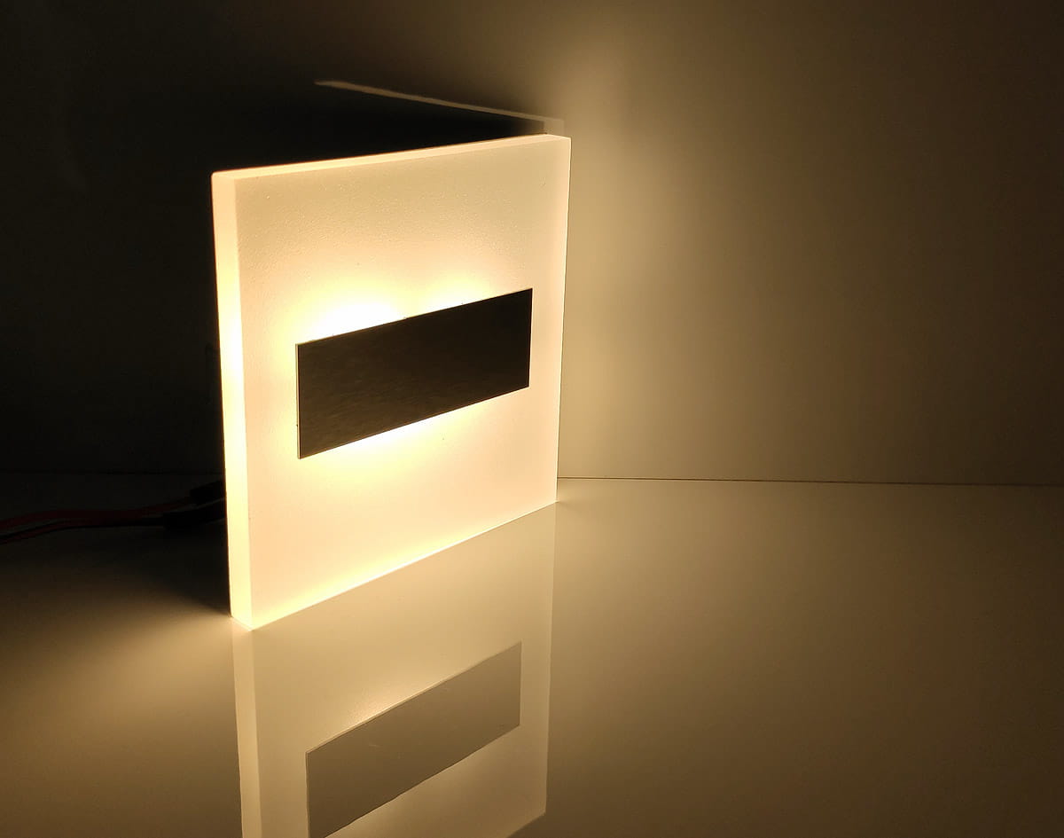 Oprawa schodowa Lumi LED 230V o ciepłej barwie światła 3000K, z matowym białym kloszem i czarnym prostokątnym elementem. Ujęcie z boku, produkt leży na płaskiej powierzchni, rzucając cień. Idealna do oświetlenia schodów.