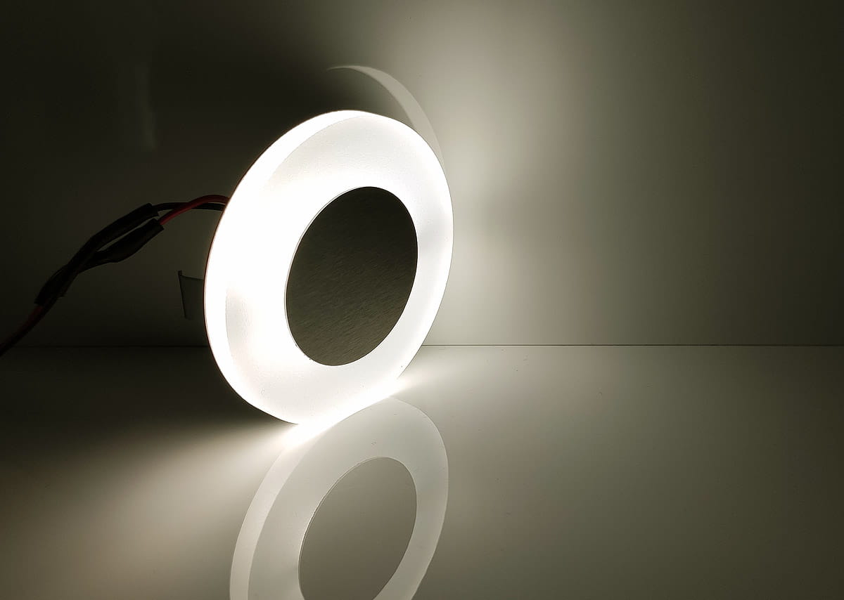 Oprawa schodowa Oti LED 230V z neutralnym światłem (4000K) o okrągłym kształcie. Wykonana z białego materiału, z widocznym czarnym wnętrzem. Produkt leży na gładkiej powierzchni, odzwierciedlając się w niej.